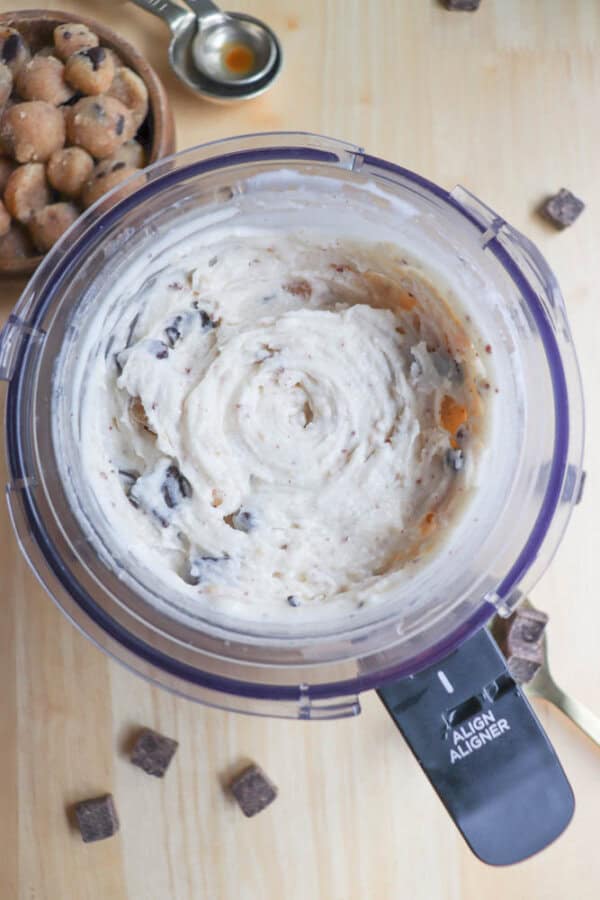 Top view of a food processor containing creamy homemade cookie dough ice cream with chocolate chunks and cookie dough pieces. A wooden bowl with more cookie dough pieces and a metal measuring spoon lie nearby on a light wooden surface.