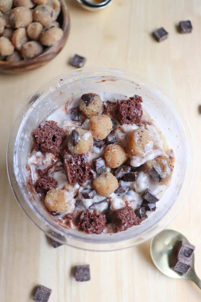 A bowl of ice cream featuring chunks of chocolate, pieces of brownie, and cookie dough balls mixed into a creamy base. Scattered chocolate pieces and cookie dough are visible on the wooden table surface.