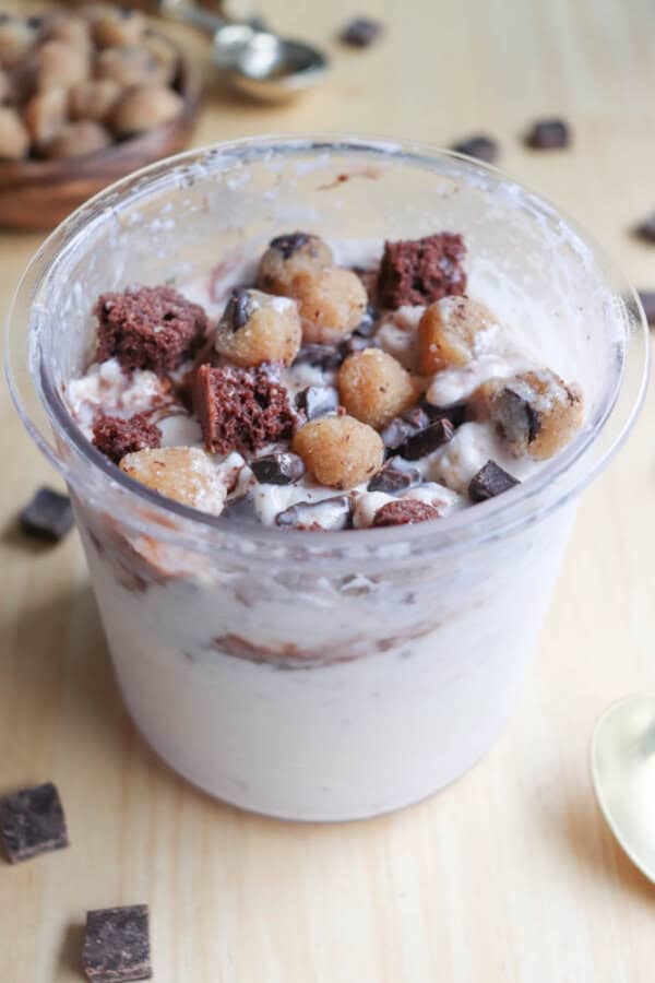 A clear cup filled with creamy ice cream mixed with chocolate chunks, brownie pieces, and cookie dough bits. A spoon and scattered chocolate pieces are in the background on a wooden surface.