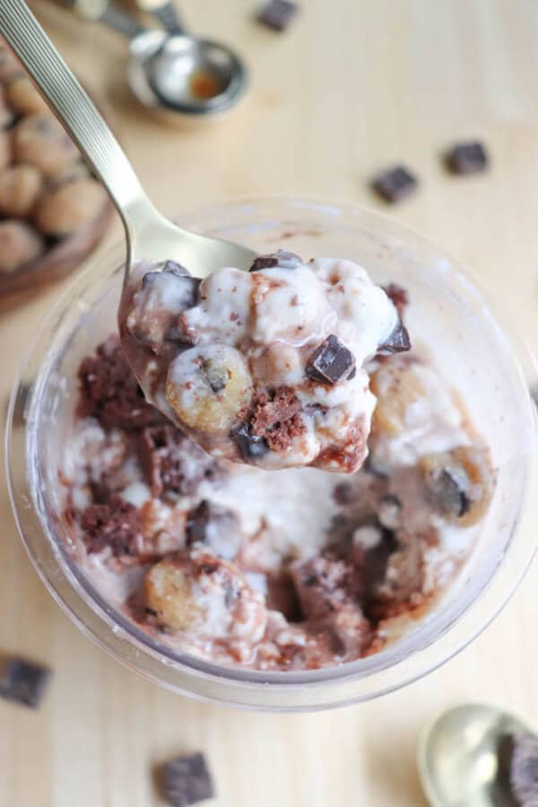 A spoonful of ice cream with chocolate chunks and cookie dough pieces is held above a clear bowl filled with more of the same dessert. The bowl rests on a wooden surface, scattered with small chocolate squares and measuring spoons.
