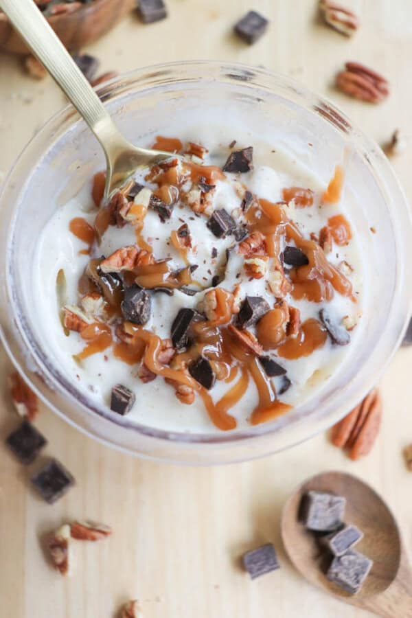 A glass bowl filled with creamy dessert topped with caramel sauce, chopped pecans, and chocolate chunks. A gold spoon rests inside the bowl. Scattered pecans and chocolate pieces are visible on the wooden surface around it.