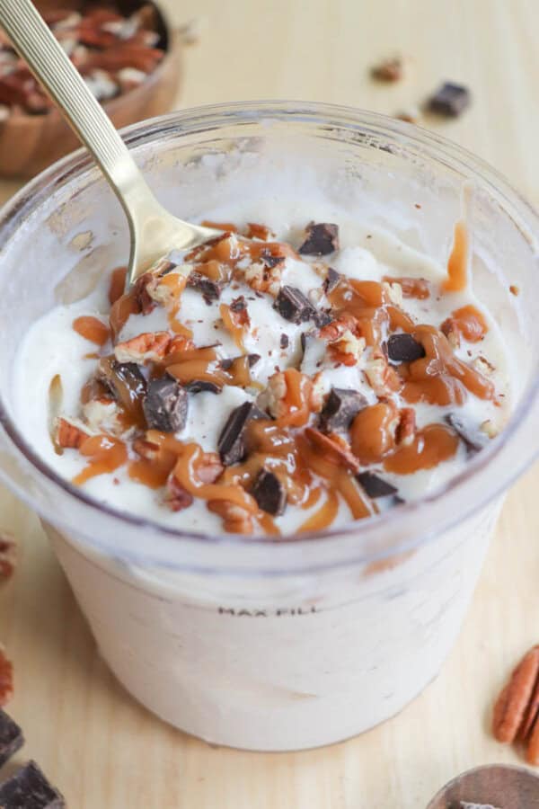 A creamy dessert in a clear cup is topped with caramel sauce, chopped nuts, and chocolate chunks. A spoon rests in the cup. A wooden surface and small bowl with additional toppings are visible in the background.