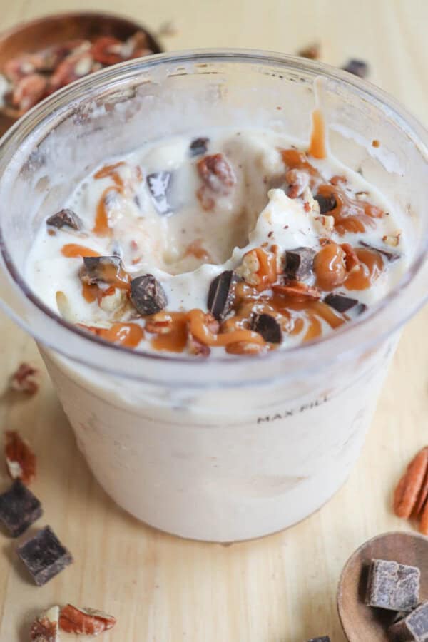 A cup filled with creamy ice cream mixed with chunks of chocolate, pecans, and swirls of caramel. The cup is on a wooden surface, surrounded by more chocolate pieces and pecans.