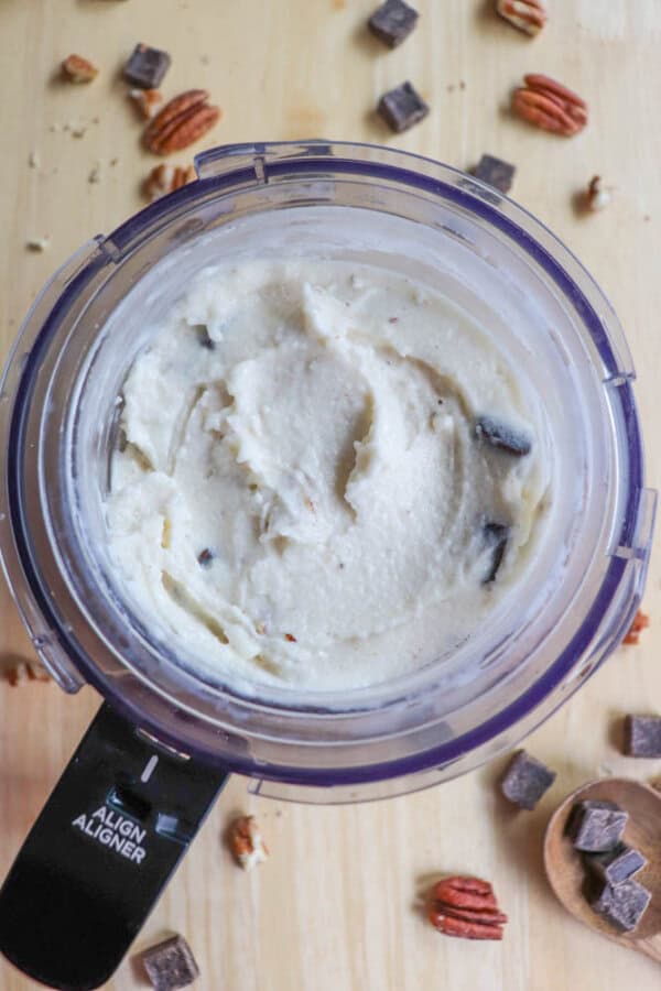 Top view of a food processor containing creamy white ice cream with visible chocolate chunks. The processor is on a wooden surface, surrounded by scattered chocolate pieces and pecans.