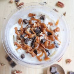 A bowl of dessert topped with whipped cream, caramel drizzle, chocolate chunks, and pecan pieces. Surrounding the bowl, more chocolate chunks and pecan pieces are scattered on a light wooden surface, with a wooden spoon nearby.