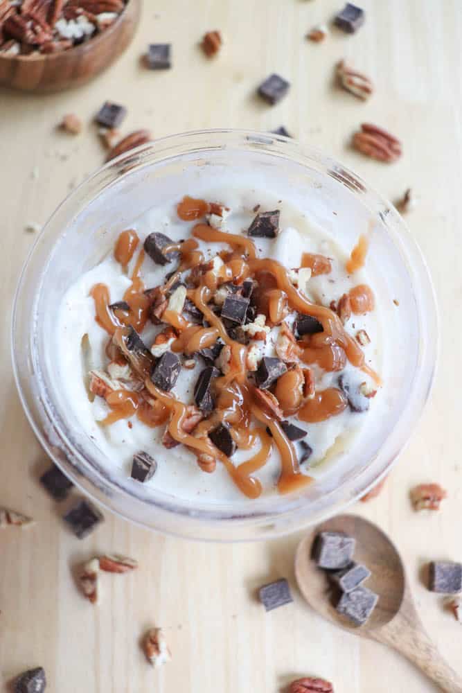 A bowl of dessert topped with whipped cream, caramel drizzle, chocolate chunks, and pecan pieces. Surrounding the bowl, more chocolate chunks and pecan pieces are scattered on a light wooden surface, with a wooden spoon nearby.