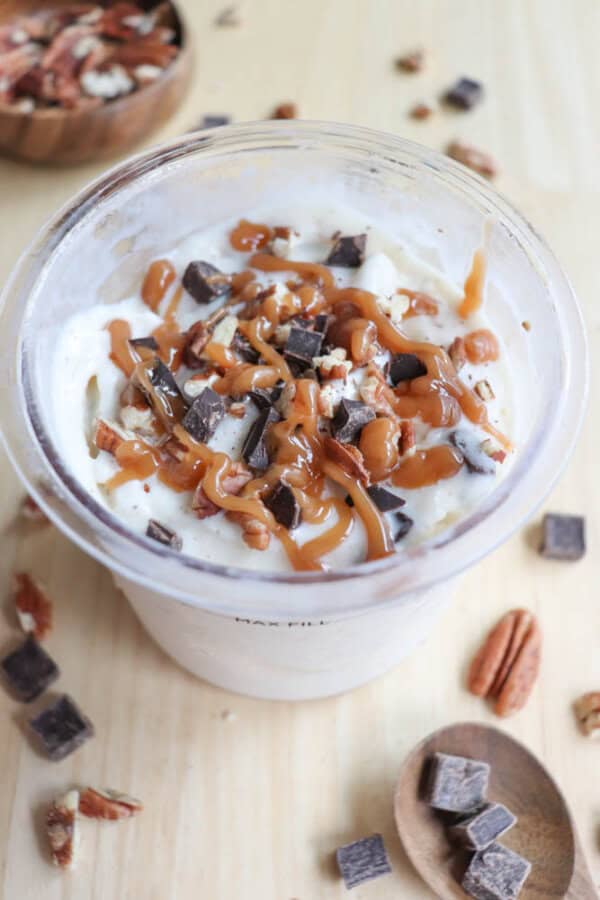 A clear cup filled with vanilla ice cream topped with caramel sauce, chocolate chunks, and chopped pecans. Wooden bowls in the background hold more chocolate pieces and pecans on a light wooden surface.