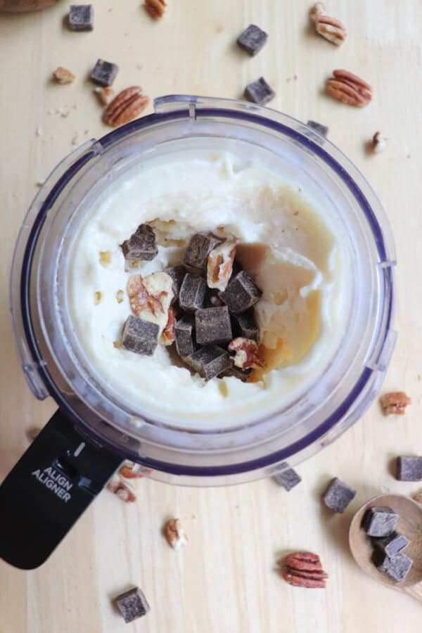 A food processor contains creamy banana ice cream topped with chunks of chocolate and pecans. Scattered around are additional chocolate pieces and pecans on a wooden surface.
