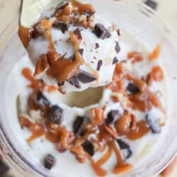 A spoon holds a scoop of creamy dessert topped with caramel sauce, chocolate chunks, and chopped nuts, over a bowl filled with the same mixture. More chocolate pieces and nuts are scattered around the wooden surface.