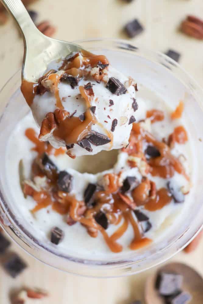 A spoon holds a scoop of creamy dessert topped with caramel sauce, chocolate chunks, and chopped nuts, over a bowl filled with the same mixture. More chocolate pieces and nuts are scattered around the wooden surface.