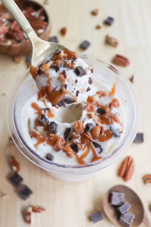 A spoonful of ice cream topped with caramel sauce, chocolate chunks, and pecan pieces is held above a container of the same dessert. Surrounding the container are additional pecans and chocolate chunks scattered on a wooden surface.