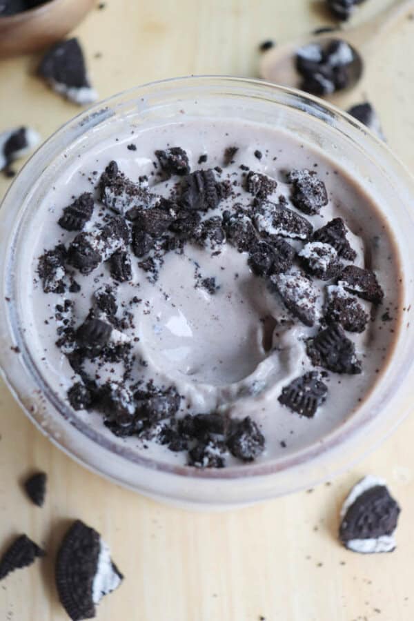 A close-up of a creamy dessert topped with crumbled chocolate cookies in a glass bowl. More cookie pieces are scattered on the light wooden surface around the bowl.