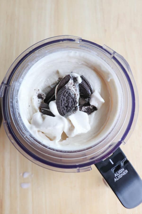 A blender filled with creamy vanilla ice cream and crumbled chocolate sandwich cookies on top, viewed from above. The blender is set on a wooden surface.