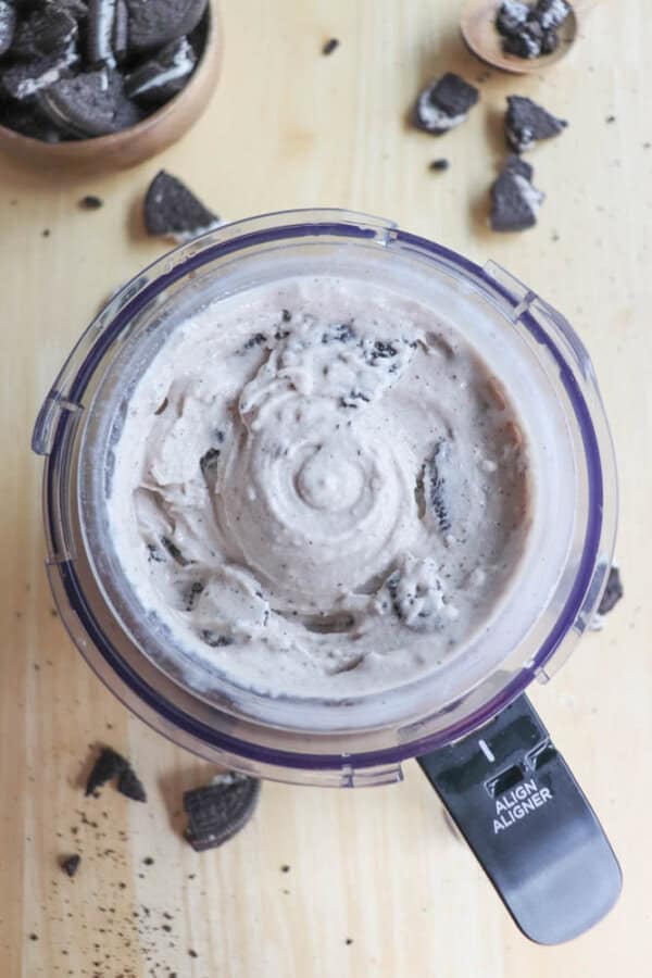 Top view of a food processor containing blended cookies and cream mixture, resembling ice cream. Cookie pieces and crumbs are scattered on the wooden surface around the processor.