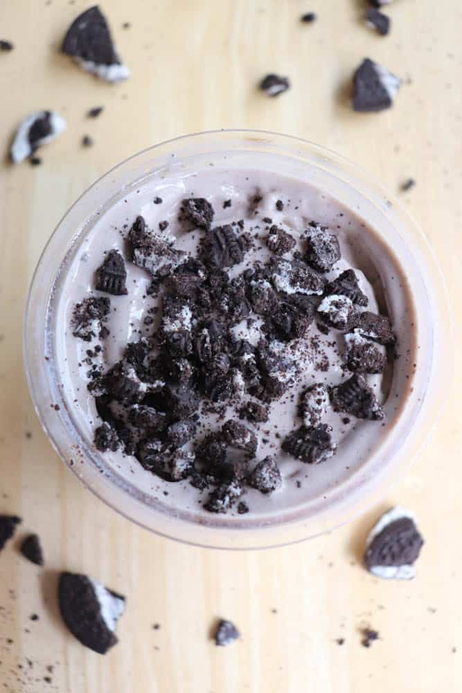 A creamy dessert in a cup topped with crushed Oreo cookies and surrounded by cookie crumbs on a light wooden surface.