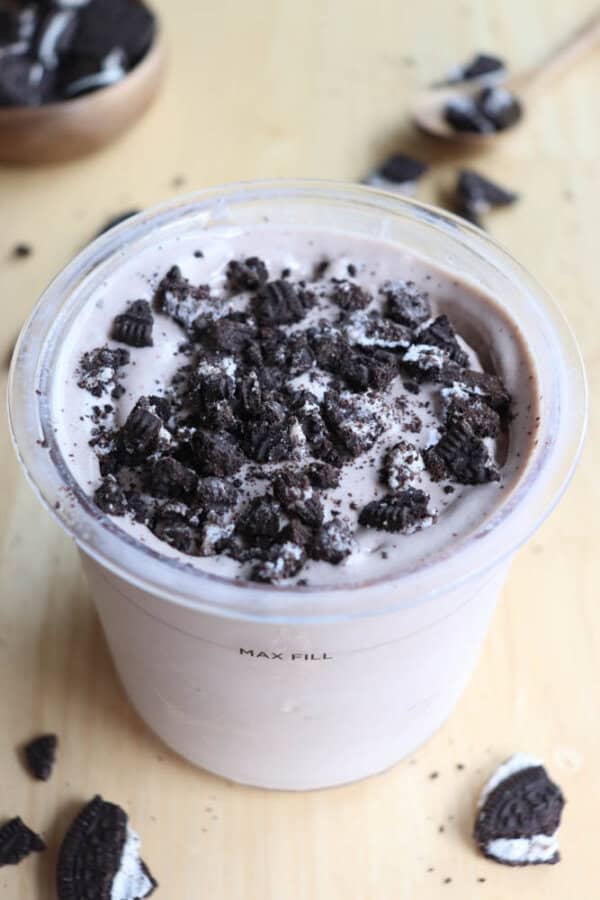 A plastic cup filled with a creamy milkshake topped with crushed chocolate cookies. More cookie pieces are scattered around the cup on a wooden surface. A wooden spoon with additional cookie bits is visible in the background.