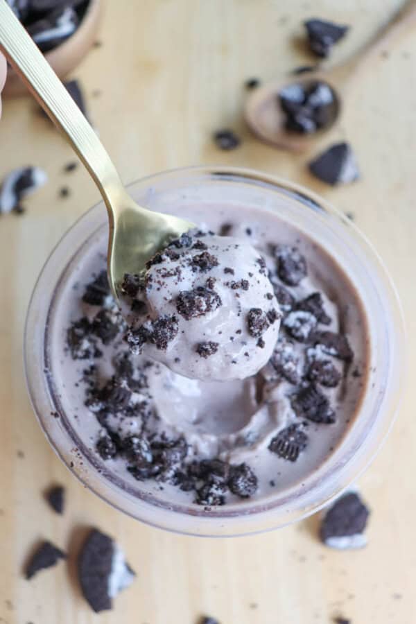 A close-up of a spoonful of cookies and cream ice cream held above a bowl. The ice cream is sprinkled with crushed chocolate cookies. Pieces of cookies and a wooden spoon are scattered on the light-colored surface.