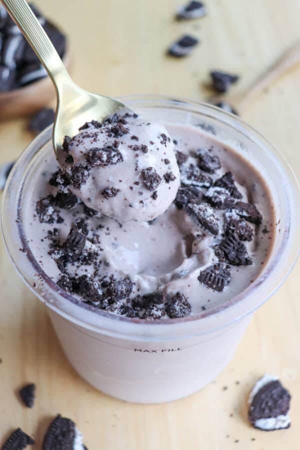 A cup of Oreo ice cream with crushed chocolate cookies on top. A spoon lifts a scoop from the cup. The ice cream is swirled and creamy, set on a light wooden surface with scattered cookie pieces around.