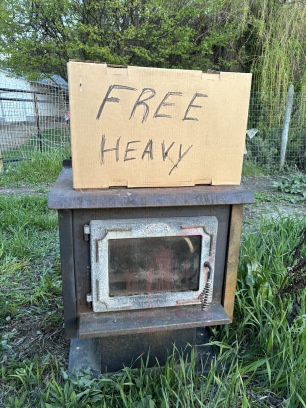 A metal wood-burning stove sits outside on grass, with a cardboard sign on top that reads FREE HEAVY in black marker. A fence and trees are visible in the background.