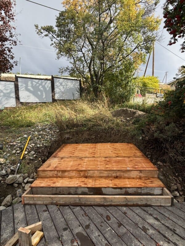 A wooden platform or deck under construction in a backyard with trees and foliage surrounding it. Theres a gravel path leading to the platform, and a yellow-handled tool rests nearby. A mix of cloudy and sunny skies is visible above.
