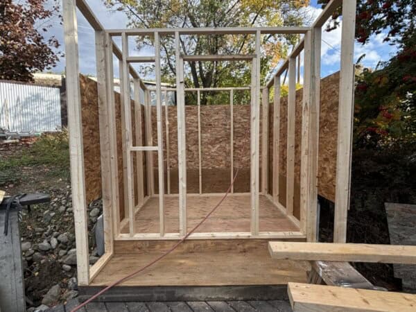 Wooden frame of a small building under construction, with a partial roof. The structure is surrounded by leafy trees and a white fence. A power cord is visible on the ground, and building materials are scattered nearby.