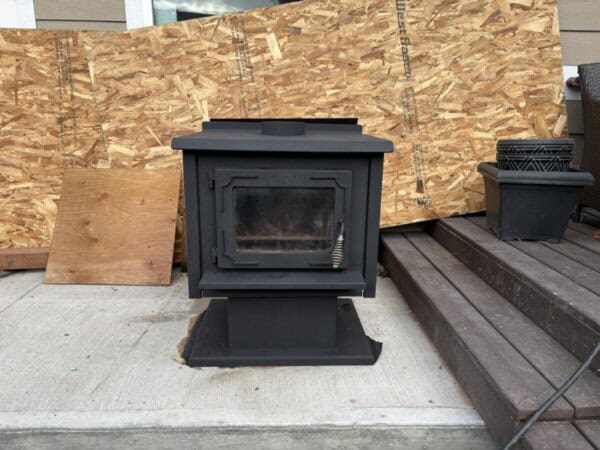A black wood-burning stove sits on a concrete surface with wooden boards as a backdrop. It has a glass front door and a coiled metal handle. A stack of dark plastic pots is visible on a wooden deck to the right.