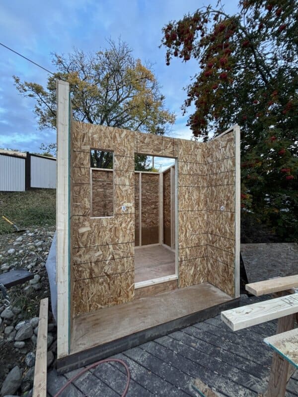 A small structure under construction with OSB board walls and openings for a door and window. The frame is set on a wooden base, surrounded by tools and materials. Trees and a cloudy sky are visible in the background.