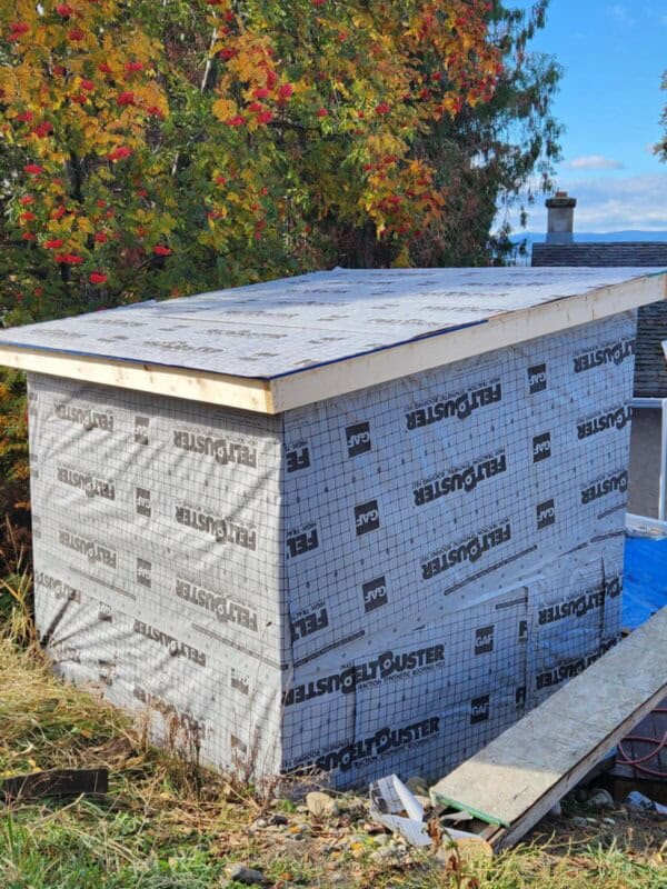 Small wooden sauna under construction with a flat roof, covered in protective house wrap labeled Weathermate and Rigidstik. Surrounded by autumn trees with colorful leaves and situated beside a house with a visible chimney in the background.