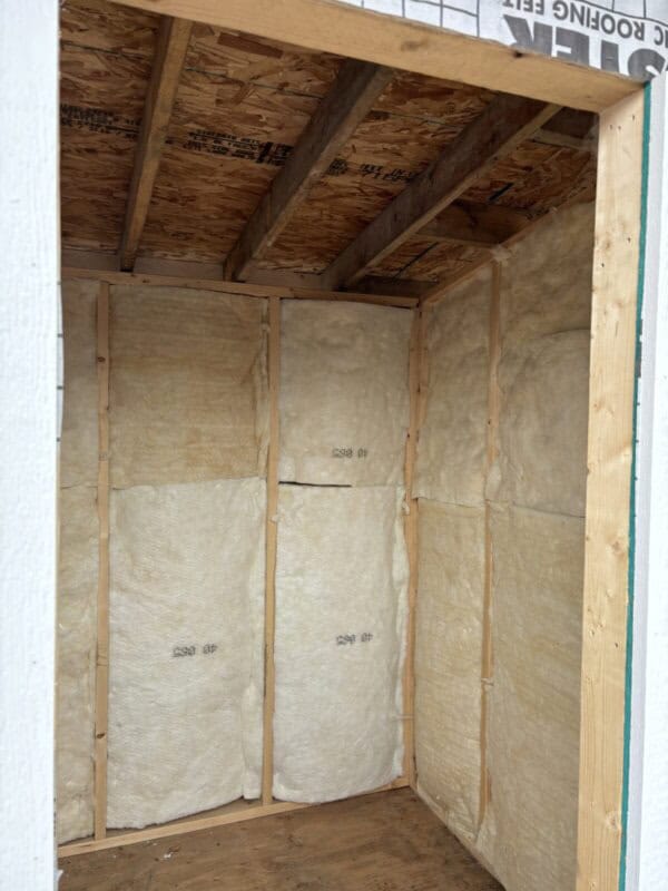 Interior view of a small sauna under construction, showing walls lined with insulation material between wooden studs. The ceiling is covered in oriented strand board (OSB). The floor is unfinished wood.