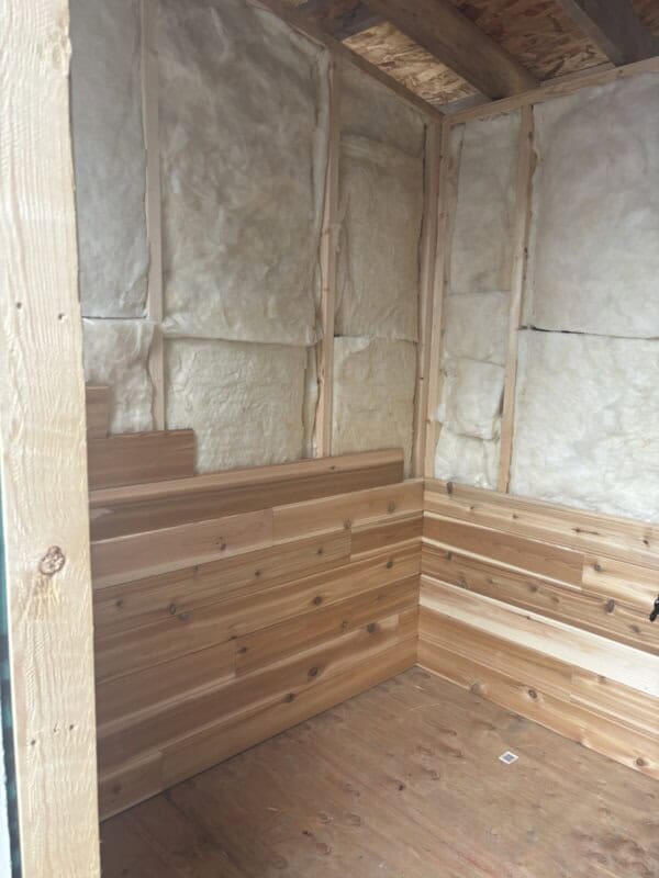 Interior view of an unfinished wooden sauna under construction, featuring partially insulated walls with fiberglass and wooden paneling on the lower half. The floor is plywood, and the ceiling beams are visible.