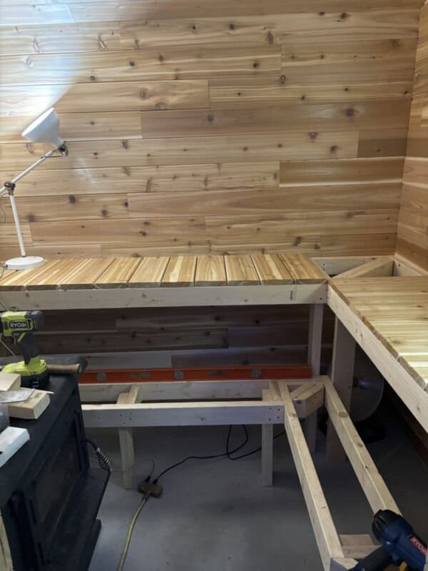 Interior view of a space with wood-paneled walls, featuring an L-shaped wooden workbench in progress. A white desk lamp is on the left, and tools like a drill and level are scattered on the workspace.