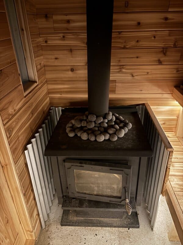 A small black stove with a metal chimney is surrounded by wooden walls. Smooth black stones are arranged on top of the stove. Metal panels cover part of the walls beside the stove. The flooring is light-colored concrete.