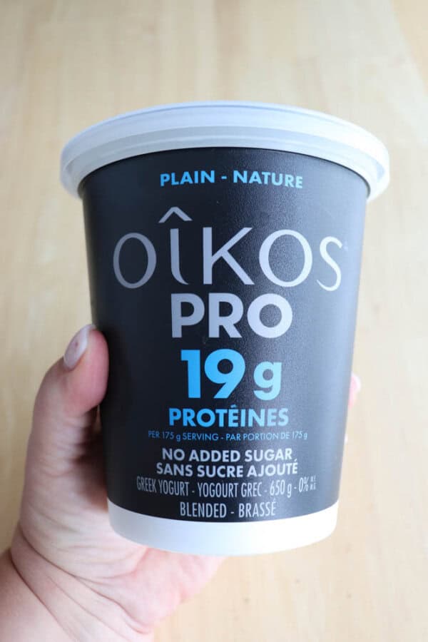 Hand holding a container of Oikos Pro Greek yogurt, plain flavor. The label states it contains 19 grams of protein per serving, with no added sugar. The container is black with white and blue text.