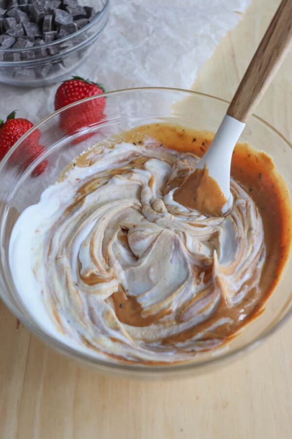 A glass bowl filled with a swirled mixture of yogurt and peanut butter, with a spatula inside. Nearby, there are two fresh strawberries and a bowl of chocolate chips on a wooden surface.
