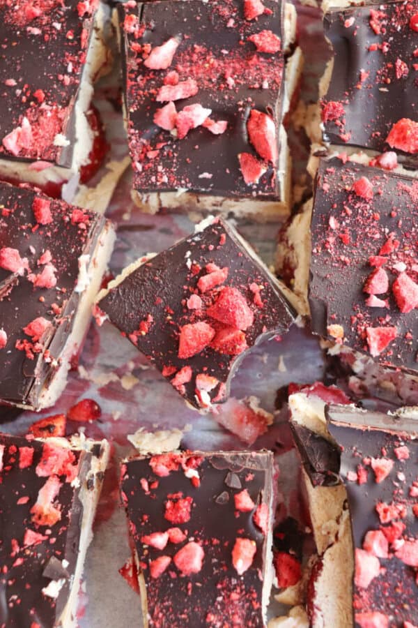 Close-up of chocolate-covered dessert bars topped with crushed red berries. One bar is bitten, revealing a creamy layer beneath the dark chocolate coating. They are arranged on a surface in a grid-like pattern.
