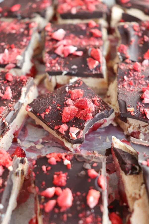 Close-up of chocolate-topped bars with a crumbly base. The bars are sprinkled with red berry pieces, giving a textured surface. The dessert is arranged on a light-colored surface.