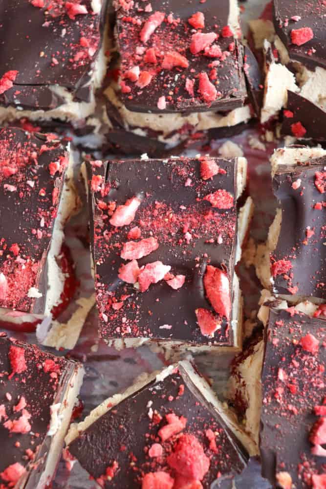 Close-up of square chocolate desserts topped with red crushed berries. The squares are arranged closely together, showcasing layers of dark chocolate and a lighter filling. The crushed berries add texture and a splash of color.