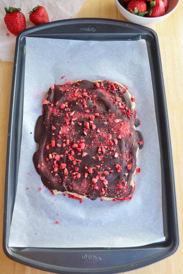 A baking tray lined with parchment paper holds a slab of strawberries and peanut butter topped with a layer of dark chocolate and sprinkled with crushed freeze-dried strawberries. A bowl of fresh strawberries is in the background.