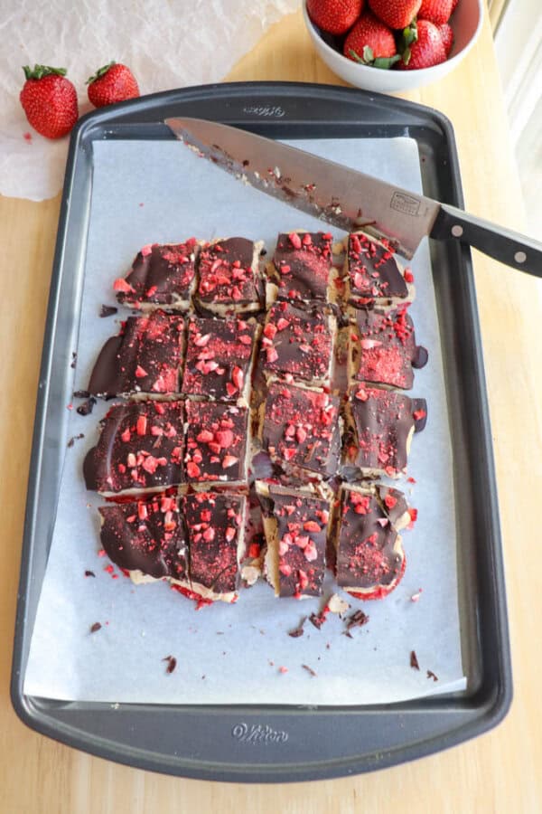 Sliced chocolate-covered squares on a parchment-lined baking tray, topped with red berry bits. A knife lies on the tray, and a bowl of strawberries is in the corner. The tray is on a wooden surface.