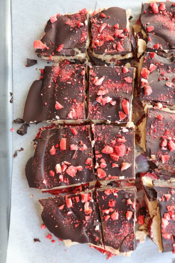 Top-down view of chocolate dessert squares on parchment paper, topped with red dried fruit pieces. Some squares have cleaner edges, while others are unevenly cut. The rich toppings contrast with the light base.