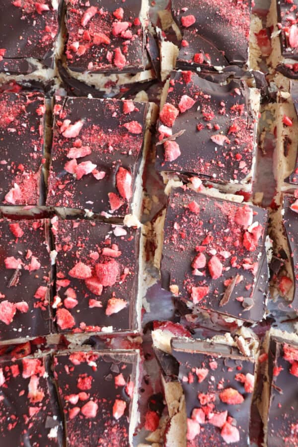 Chocolate-topped dessert squares sprinkled with red crushed pieces, possibly berries, arranged in a grid pattern. The top layer of chocolate appears glossy, with bits of red contrasting against the dark surface.