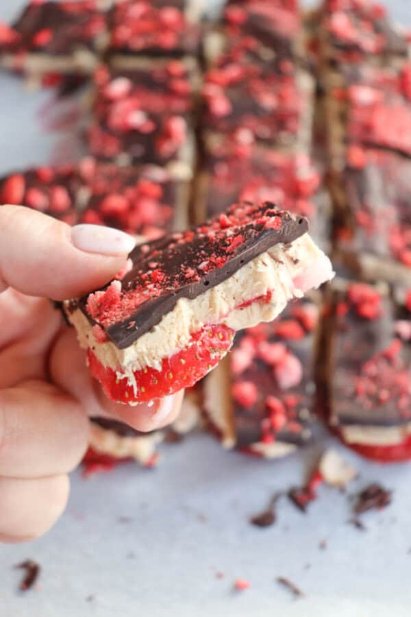 A hand holds up a treat composed of layers of chocolate, creamy filling, and red fruit topping, with more treats blurred in the background. The layered dessert appears to have a crunchy texture with visible red fruit pieces.