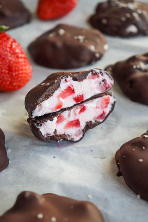 Chocolate-covered strawberry bites are displayed. One bite is halved to reveal creamy filling with strawberry pieces. A whole strawberry is seen in the background, with all items on a parchment-lined surface.