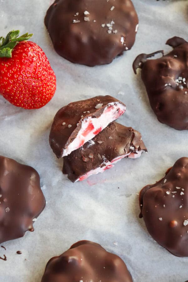 Chocolate-covered strawberries filled with cream sit on parchment paper. One is cut open to reveal the creamy filling with strawberry pieces inside. A fresh strawberry is placed alongside the chocolates, which have a slight dusting of salt or sugar.