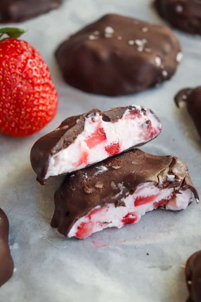 Close-up of chocolate-covered strawberry yogurt bites with two halves showing the creamy yogurt and strawberry inside. A fresh strawberry is nearby, and other chocolate-covered bites are in the background on a parchment paper-lined surface.