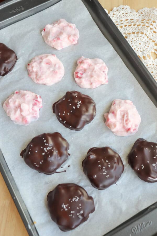 A baking tray lined with parchment paper holds clusters of chocolate-covered and pink, creamy clusters. The chocolate ones are sprinkled with a hint of sea salt. A lace doily peeks from the corner of the wooden table.