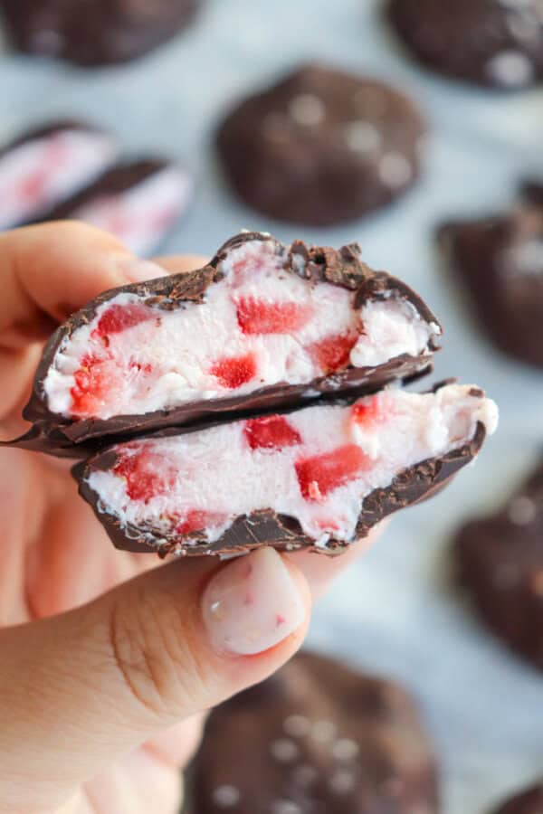 Close-up of a hand holding two halves of a chocolate-covered treat, revealing a creamy white filling with red strawberry pieces inside. Other similar treats are blurred in the background.
