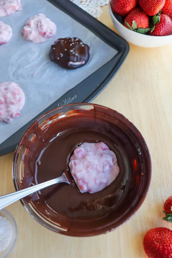 A spoon dips a strawberry and cream mixture into a bowl of melted chocolate. A baking sheet with more strawberry treats and a bowl of fresh strawberries are nearby on a wooden surface.