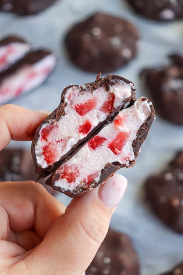 A hand holds a halved chocolate-coated treat filled with yogurt and pieces of red strawberry. Blurred background shows more similar treats on a parchment-lined surface.