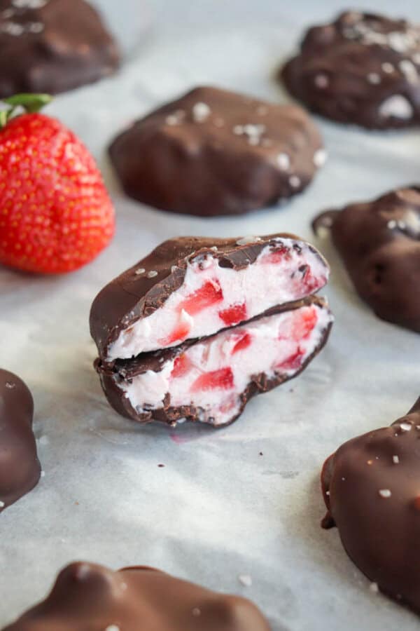 Chocolate-covered strawberry yogurt bites are scattered on parchment paper. One bite is cut open, revealing a creamy white and pink filling with strawberry pieces. A fresh strawberry is in the background.